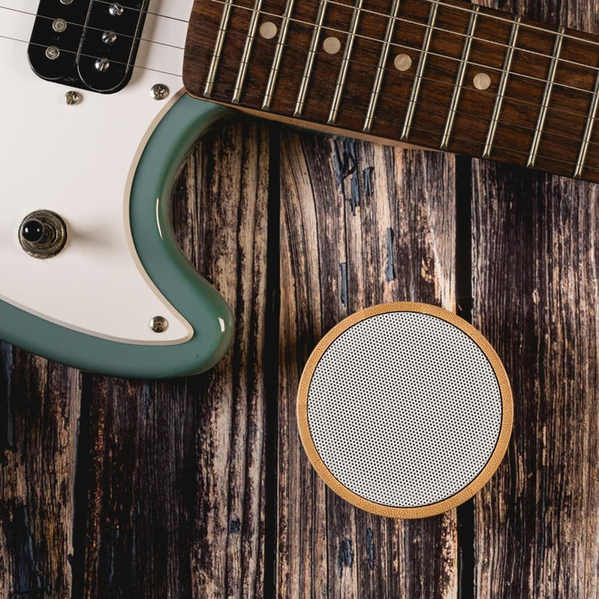 A round white Bluetooth speaker with a wooden base, placed beside a teal electric guitar on a rustic wooden surface.