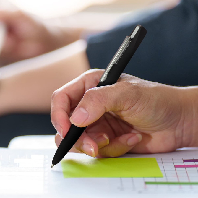 A sleek black metal pen is held in a hand, writing on a piece of paper with a bright sticky note.
