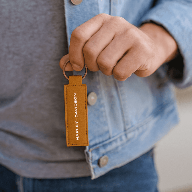 A tan recycled leather keyring held by a person wearing a denim jacket, featuring branding.