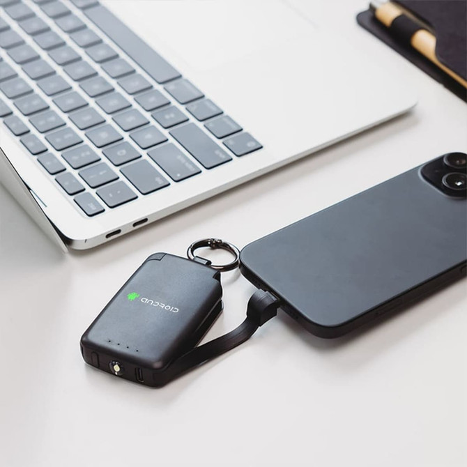 A black 2,000mAh mini power bank with a keyring attached, connected to a smartphone, placed next to a laptop.