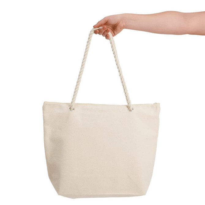 A hand holds a large, cream-coloured canvas tote bag with rope handles. The bag is plain and unbranded.