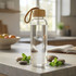 A clear glass drink bottle with a wooden cap, displayed on a countertop with mint leaves and smooth stones.