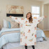 A cosy, cream-coloured wearable blanket featuring playful animal designs, displayed in a bedroom setting.