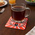 A drink bottle sits on a red cork coaster featuring a graphic design, on a wooden table with food and drink items.