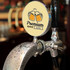 A shiny silver beer tap with a circular badge featuring two mugs of beer, set against a blurred bar background.