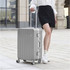 A silver ribbed ABS suitcase with wheels stands on a tiled surface next to a person wearing a skirt and sneakers.