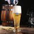 A tall, clear glass filled with pale beer and a frothy head, displayed alongside a wooden barrel and a lantern.