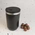 A reusable black coffee cup with a stainless steel band and a black lid, alongside a handful of chocolate balls.