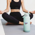 A drink bottle in mint green with a lid and a logo, placed on a yoga mat beside a person in activewear.