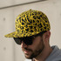 A flat peak cap featuring a yellow and black abstract design, worn by a person in sunglasses.