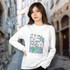 A woman in a white long sleeve crew shirt with a floral graphic design, standing in a cobblestone street setting.
