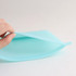 A teal silicone reusable storage pouch with a smooth surface and a hand holding it. Features a logo.