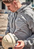 A man in a light grey unisex rain jacket holds a safety helmet, with a construction site in the background.
