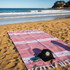 A pink and white striped Turkish towel laid out on the sand, accompanied by a black hat and a camera.