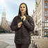 A woman wearing a black puffer jacket stands in an outdoor urban setting with historic buildings in the background.