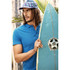 A man in a blue polo shirt holds a surfboard. He wears a patterned cap and stands near palm leaves.