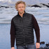 A man stands by a glacial lake wearing a black thermal vest over a long-sleeve top, with mountains in the background.