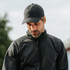 A black cap worn by a man in a black jacket, with a blurred outdoor background. The cap features a logo.