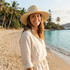 A straw hat in natural tones, worn by a woman on a beach, surrounded by palm trees and water.