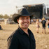 A man wearing a black straw hat smiles, with an outdoor festival stage and crowd in the background.