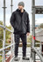A man wearing a black unisex softshell jacket stands on scaffolding, paired with black trousers and boots.