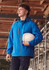 A man wears a blue unisex packable rain jacket, holding a white helmet, set against a construction backdrop.