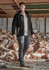A model wearing black work pants and a black fleece jacket stands outdoors, with stacks of timber in the background.
