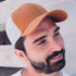 A cap featuring a brown leather front and grey fabric sides, worn by a man with a beard against a concrete background.