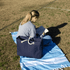 A navy waterproof beach bag with rope handles rests beside a person reading on a blue and white blanket.