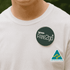 A 90mm button badge on a white shirt, featuring a dark green background and a logo.