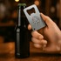 A drink bottle opener resembling a credit card, made of stainless steel, held in a hand next to a dark bottle.