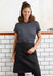 A woman wearing a black short waisted apron over a grey t-shirt, standing against a tiled wall. The apron features a logo.