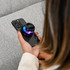 A compact, black speaker with colourful lights rests on a smartphone in a person's hand. The background features a textured fabric.