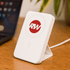 A white wireless charging power bank with a logo, positioned on a wooden surface next to a laptop.