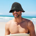 A man wearing a black bucket hat and sunglasses, sitting on a beach with ocean waves in the background.