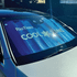 A foldable car sunshade in blue with branding, placed on the windshield of a silver car.