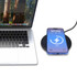A black wireless charger with a smartphone displaying a charging indicator, beside a laptop with a natural backdrop.