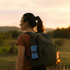 A woman with a ponytail wearing a brown top and carrying a green backpack, set against a sunset landscape.