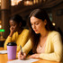 A powder coated stainless steel coffee mug in lavender sits on a table beside two women studying. The mug has a black lid.