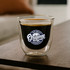 A double wall glass featuring a black beverage and a logo, set on a wooden table. The glass has a transparent design.