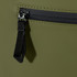 A close-up of a dark olive green roll-top backpack featuring a zippered pocket and a black zipper pull.