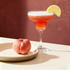 A peach-coloured margarita glass filled with a frothy drink and garnished with a lemon slice, next to a fresh peach on a plate.