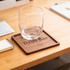 A square wooden coaster in dark brown with a glass on top, placed on a wooden desk next to a notepad.