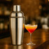 A stainless steel cocktail shaker in a polished finish, next to a vibrant cocktail in a glass.