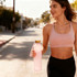 A drink bottle in a light pink colour with a gold cap, held by a person in activewear on a street.
