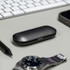 A black charging case for true wireless earbuds rests on a desk next to a watch, a smartphone, and a pen.