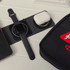 A black tech kit featuring a smartwatch, wireless earbuds case, and space for a smartphone. Includes a branded logo.