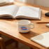 A white ceramic mug with a handle, placed on a wooden table, alongside notebooks and an open book.