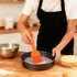 An orange pastry brush is being used to apply something in a round baking tin, with a mixing bowl and cake nearby.