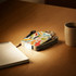 A mini power bank with a colourful design and a flashlight feature on a wooden table beside a mug and notepad.